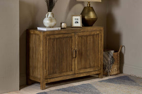 The Yasha Reeded Mango Wood Sideboard by nkuku, with vertical grooves, stands against a beige wall. On top are a vase with dried flowers, a book, candle, framed photo, and brass lamp. A woven basket sits beside a blue and beige rug.