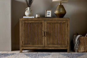 The nkuku Yasha Reeded Mango Wood Sideboard stands against a beige wall, topped with a ceramic vase of dried flowers, candle, framed photo, and gold lamp. A woven basket sits on the floor nearby, with part of a rug visible.