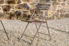 A brown nkuku Yealme Iron Bistro Chair with a slatted seat and backrest stands on gravel before a stone wall of irregular brown and tan rocks. Another piece of industrial outdoor furniture is partially visible at the left edge.