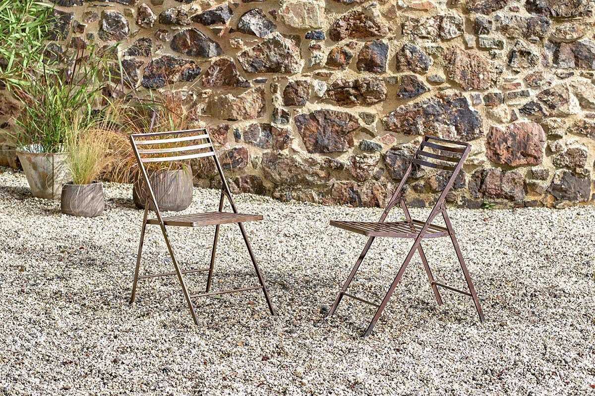 Two brown nkuku Yealme Iron Bistro Chairs face each other on a gravel patio, set against a rustic stone wall and surrounded by potted plants, creating a relaxed spot to enjoy industrial outdoor furniture in the fresh air.