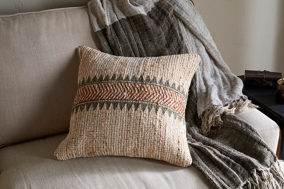 A textured beige couch features the nkuku Zairya Jute & Cotton Cushion Cover in natural and rust, adding warmth with its earthy tones and woven patterns.