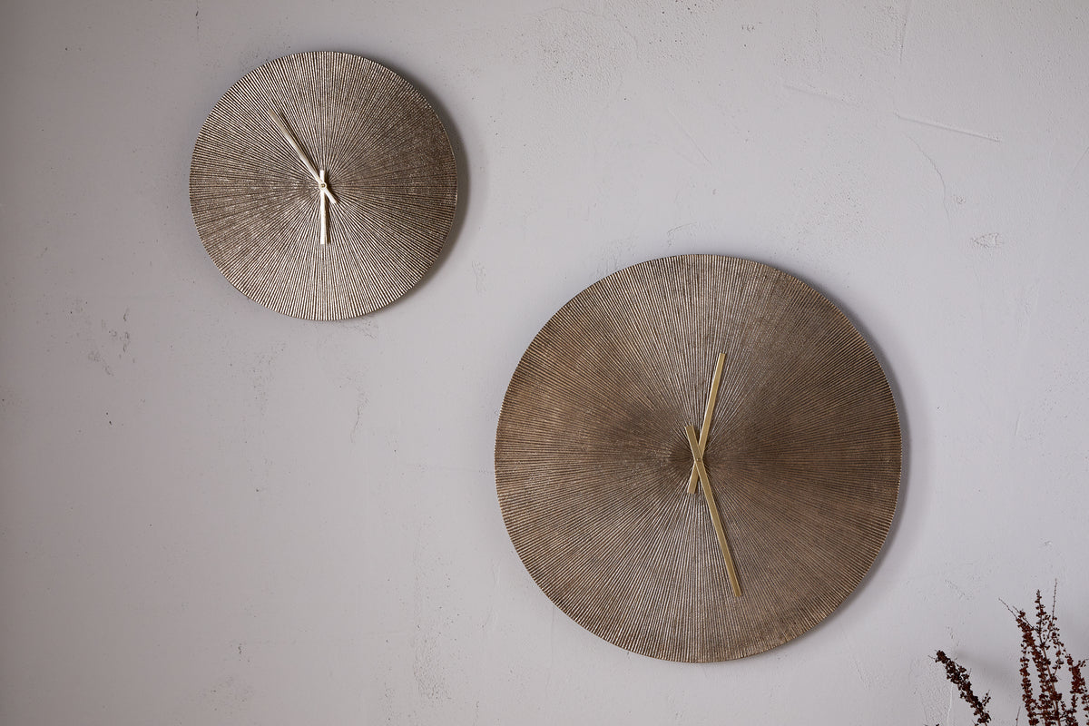 Two Zuwena Etched Wall Clocks by nkuku in antique brass are mounted on a light textured wall. The minimalist display features different sizes, with dried plant stems in the lower right corner adding subtle decor.