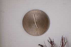 A round, textured Zuwena Etched Wall Clock in antique brass by nkuku, with no numerals and hands at 12:00, hangs on a light gray wall. Dried brown foliage in the corner adds a natural touch to this minimalist display.