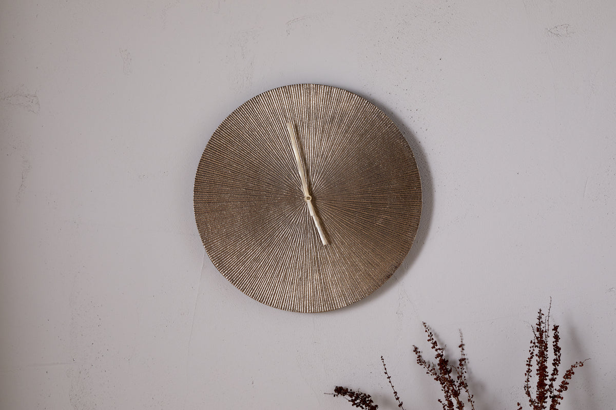 A round, textured Zuwena Etched Wall Clock in antique brass by nkuku, with no numerals and hands at 12:00, hangs on a light gray wall. Dried brown foliage in the corner adds a natural touch to this minimalist display.