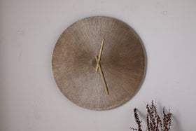 The nkuku Zuwena Etched Wall Clock in Antique Brass, featuring a textured radial gold face and minimal gold hands, hangs on a plain light wall with dried brown plant stems in the corner for a minimalist accent.