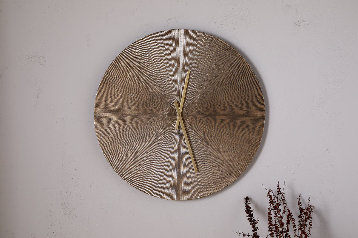 The nkuku Zuwena Etched Wall Clock in Antique Brass, featuring a textured radial gold face and minimal gold hands, hangs on a plain light wall with dried brown plant stems in the corner for a minimalist accent.