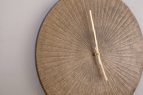 The nkuku Zuwena Etched Wall Clock in Antique Brass features a textured, round face with a radial beige pattern and slim pale gold hands. This minimalist, unmarked clock adds sophistication when mounted on a light gray wall, displaying 12:25.
