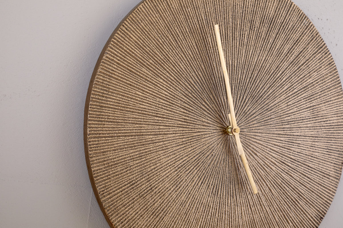 The nkuku Zuwena Etched Wall Clock in Antique Brass features a textured, round face with a radial beige pattern and slim pale gold hands. This minimalist, unmarked clock adds sophistication when mounted on a light gray wall, displaying 12:25.