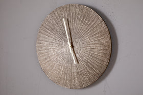 The Zuwena Etched Wall Clock by nkuku features a radial, antique brass finish and slim silver hands set to 11:00. This minimalist piece has no numbers and is mounted on a plain light gray wall.