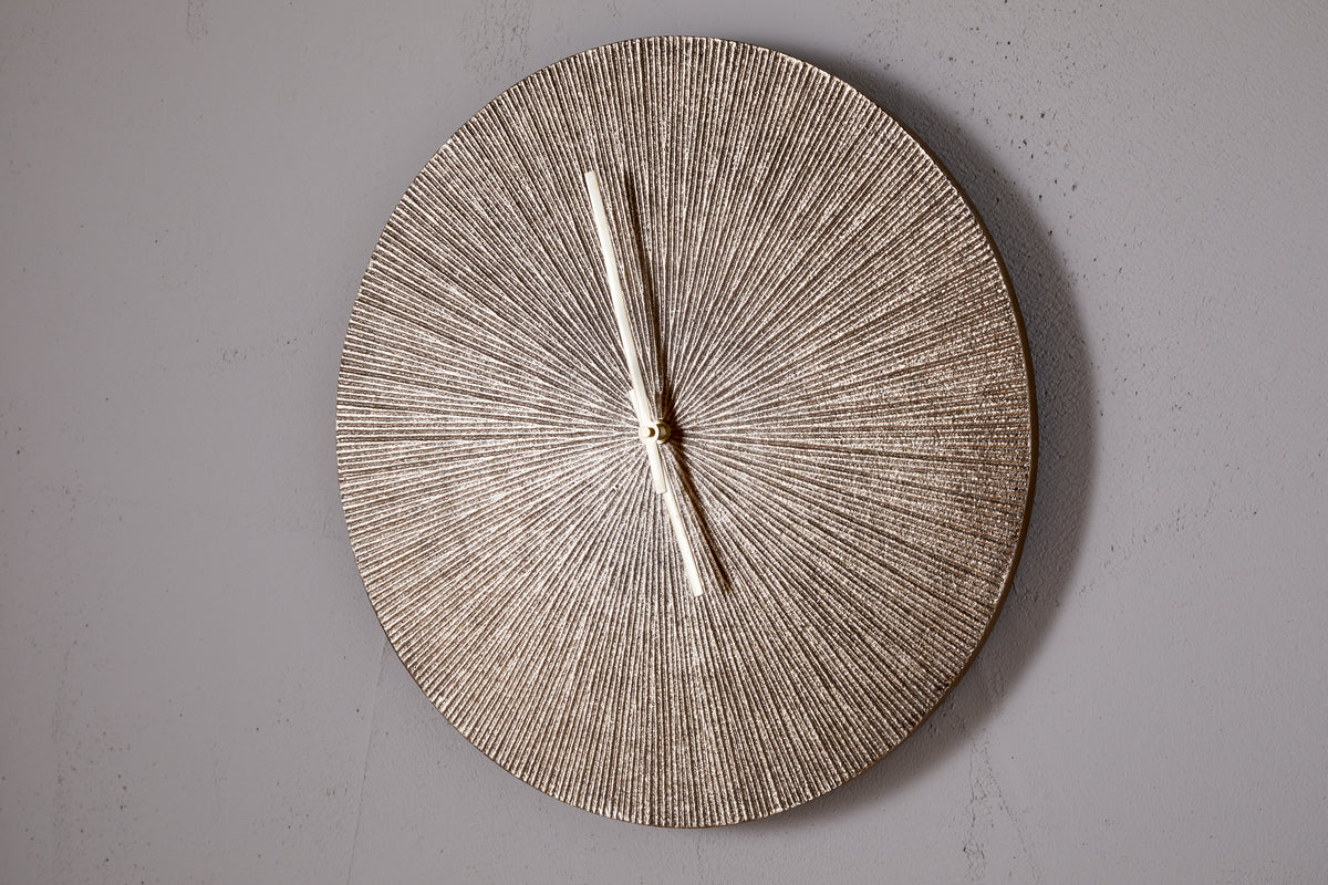 The Zuwena Etched Wall Clock by nkuku features a radial, antique brass finish and slim silver hands set to 11:00. This minimalist piece has no numbers and is mounted on a plain light gray wall.