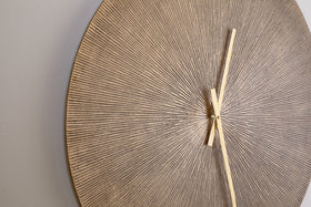 A close-up of the nkuku Zuwena Etched Wall Clock - Antique Brass shows its round, textured face with a beige and gold radial pattern. Minimalist gold hands point to 1:25 against a light gray background, with part of the clock face visible.