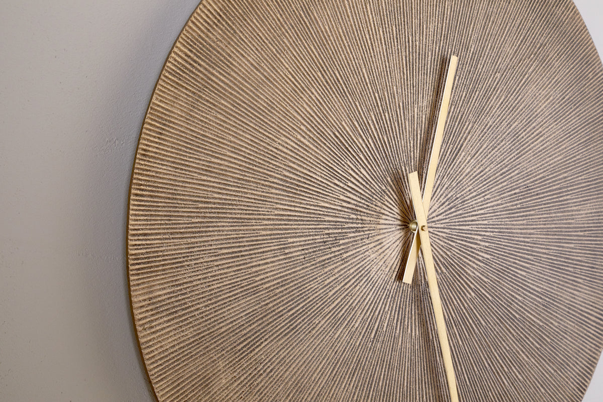 A close-up of the nkuku Zuwena Etched Wall Clock - Antique Brass shows its round, textured face with a beige and gold radial pattern. Minimalist gold hands point to 1:25 against a light gray background, with part of the clock face visible.