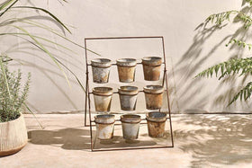 The nkuku Abari Planter Stand displays nine aged zinc pots in a 3x3 grid. Set on concrete by a beige wall, it's framed by leafy plants and gentle shadows, bringing rustic garden style and sunlit charm to any space.