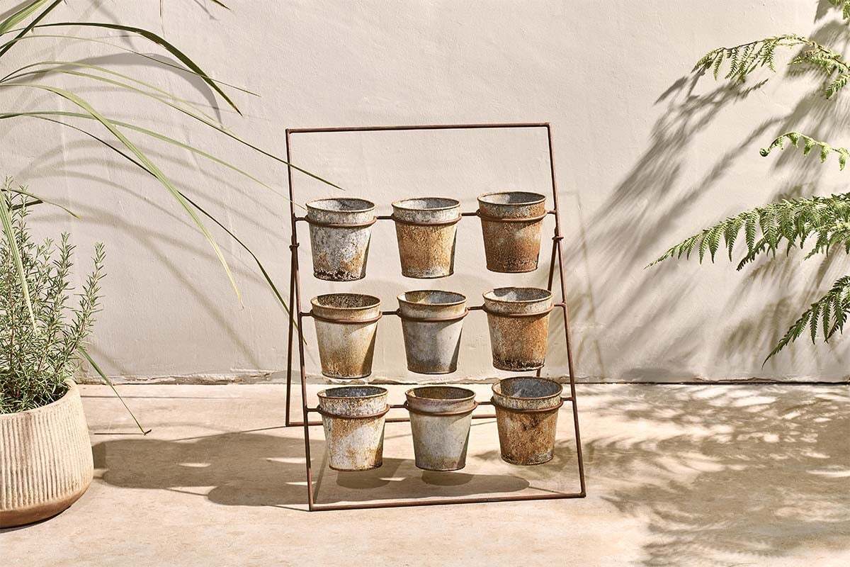 The nkuku Abari Planter Stand displays nine aged zinc pots in a 3x3 grid. Set on concrete by a beige wall, it's framed by leafy plants and gentle shadows, bringing rustic garden style and sunlit charm to any space.