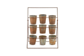 The Abari Planter Stand by nkuku holds nine rustic aged zinc pots in three rows. Angled backward for display, this minimalist metal rack showcases weathered pots—ideal as unique garden decor—against a plain white background.