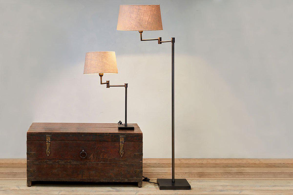 A Kara Iron Floor Lamp - Bronze by nkuku stands on the wooden floor near a pale wall, beside a matching table lamp with beige or jute shade and black or aged bronze base; the smaller rests atop an old wooden trunk.