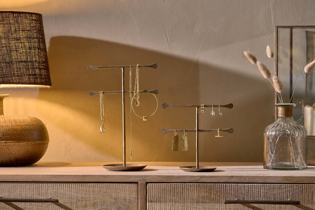 Two nkuku Liman Jewellery Stands, each displaying necklaces, bracelets, and earrings, are on a wooden dresser. A textured lamp with a brown shade is to the left, and a glass vase with dried plants sits to the right against a beige wall.