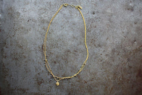The Mai Necklace by nkuku features a delicate, gold-toned asymmetric design with two fine chains—one with small beads and the other with a round pendant—plus a lobster clasp and branded tag near the closure. Photographed on textured gray.