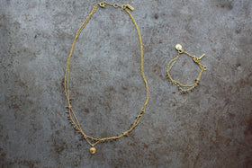 The nkuku Mai Necklace, featuring two gold chains with small beads, pyrite stones, and a round pendant, is shown on a textured gray surface beside a matching bracelet with pyrite beads and a charm-accented clasp.
