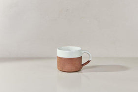 The Mali Ribbed Coffee Mug - White (Set of 2) by nkuku features a handmade design with a white upper half, ribbed terracotta lower half, and a white handle, displayed on a light beige surface against a pale background.