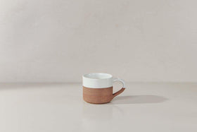 The Mali Ribbed Espresso Mug - White by nkuku, featuring a ribbed terracotta base, rests on a light surface against a neutral wall.
