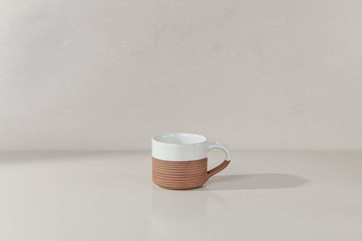 The Mali Ribbed Espresso Mug - White by nkuku, featuring a ribbed terracotta base, rests on a light surface against a neutral wall.