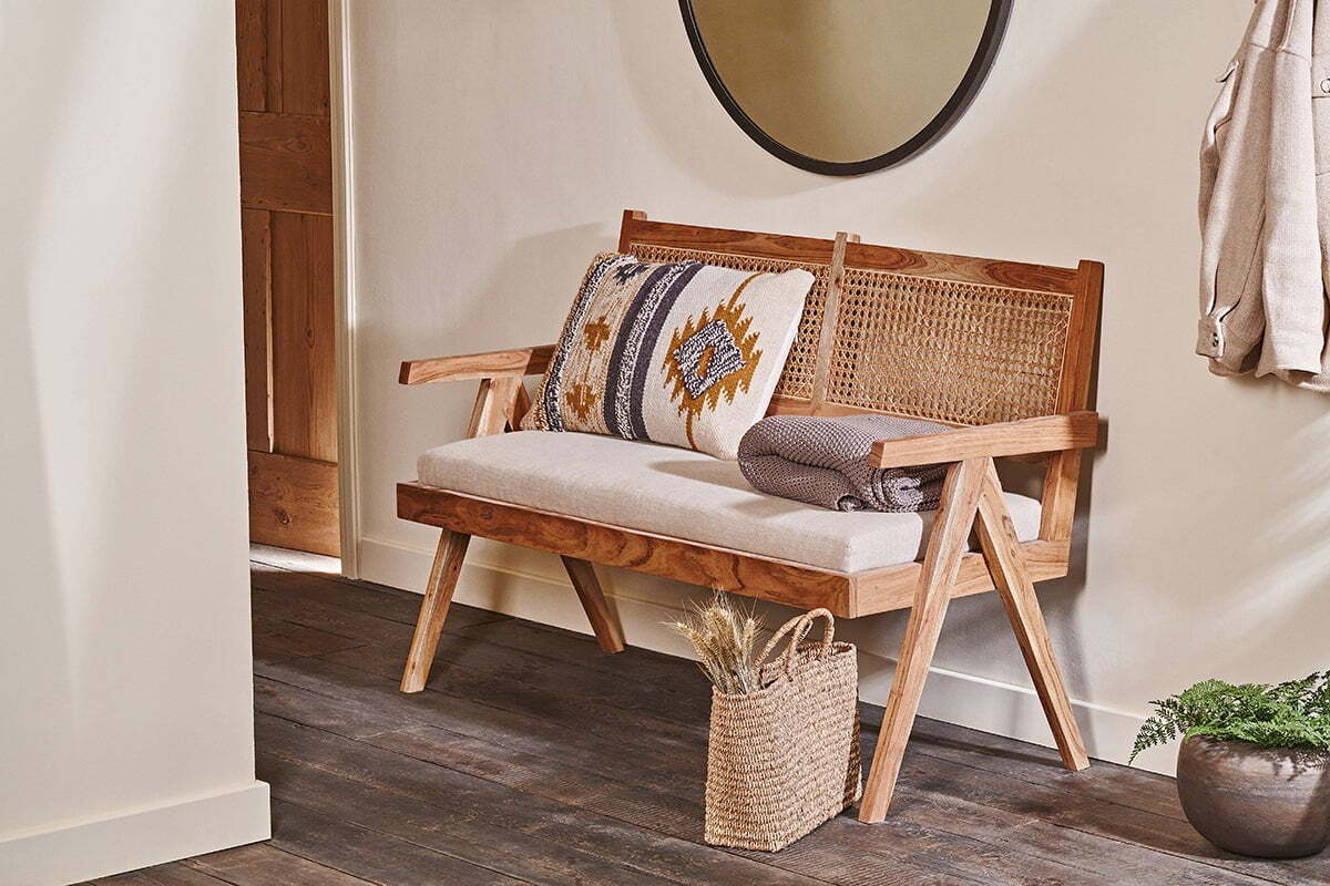 The nkuku Nalrina acacia and cane sofa with a beige linen seat pad, patterned pillows, and folded brown blanket sits against a cream wall below a round mirror. A basket of dried grasses rests on the dark wood floor nearby.
