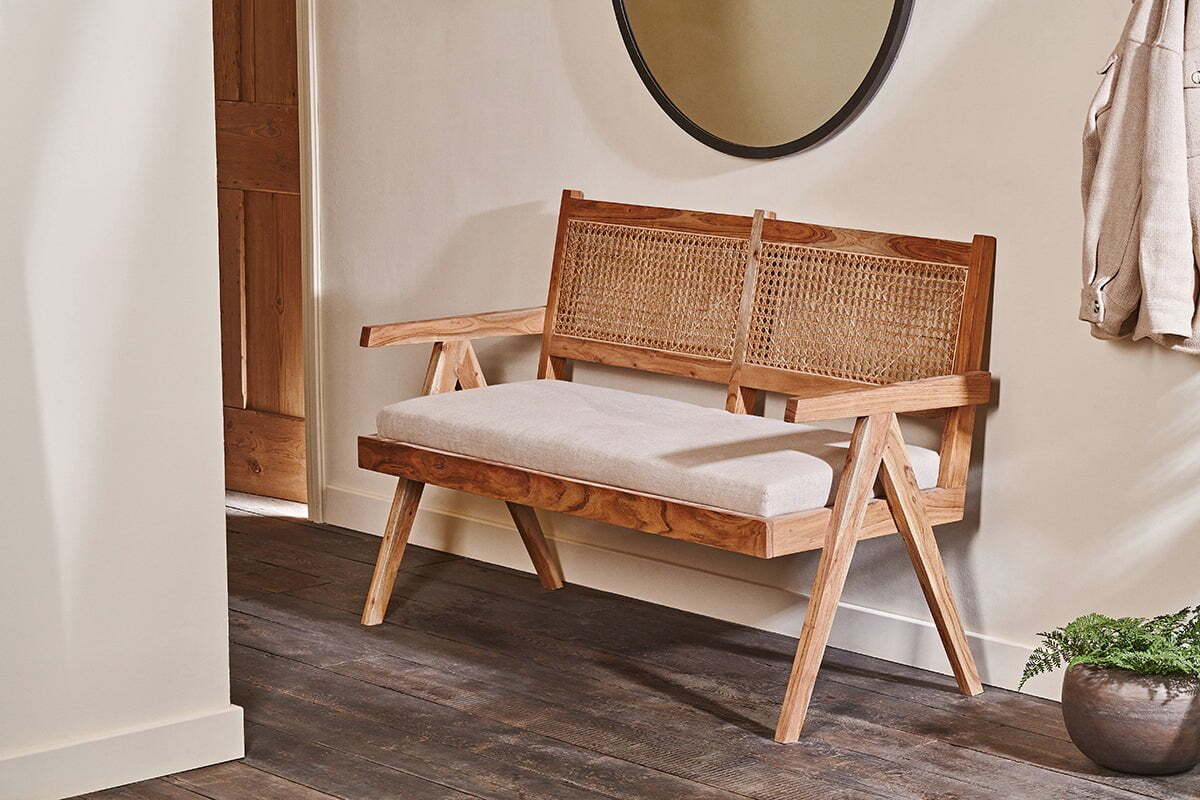 The nkuku Nalrina acacia and cane sofa with a light cushion sits on a dark wood floor, with a mirror hanging on the wall behind it.