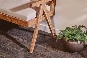The Nalrina acacia and cane sofa by nkuku rests on a dark wood floor beside a potted fern, featuring a beige linen seat pad.
