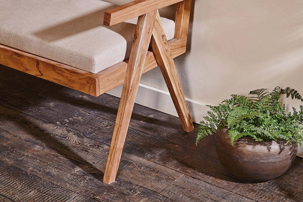 The Nalrina acacia and cane sofa by nkuku rests on a dark wood floor beside a potted fern, featuring a beige linen seat pad.