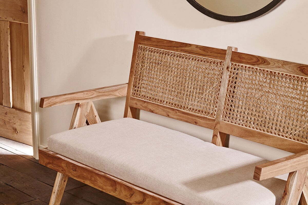 The Nalrina acacia and cane sofa by nkuku features natural acacia wood, a woven cane backrest, and a light beige cushioned seat, blending classic style with eco-friendly design against a cream wall.