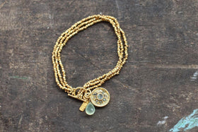 The Pandita Charm Bracelet by nkuku is a handmade piece with three strands of small gold-plated beads. It features an ornate medallion, a pale green teardrop stone, and a gold bar charm, set on a dark wooden surface.