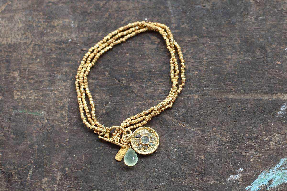 The Pandita Charm Bracelet by nkuku is a handmade piece with three strands of small gold-plated beads. It features an ornate medallion, a pale green teardrop stone, and a gold bar charm, set on a dark wooden surface.