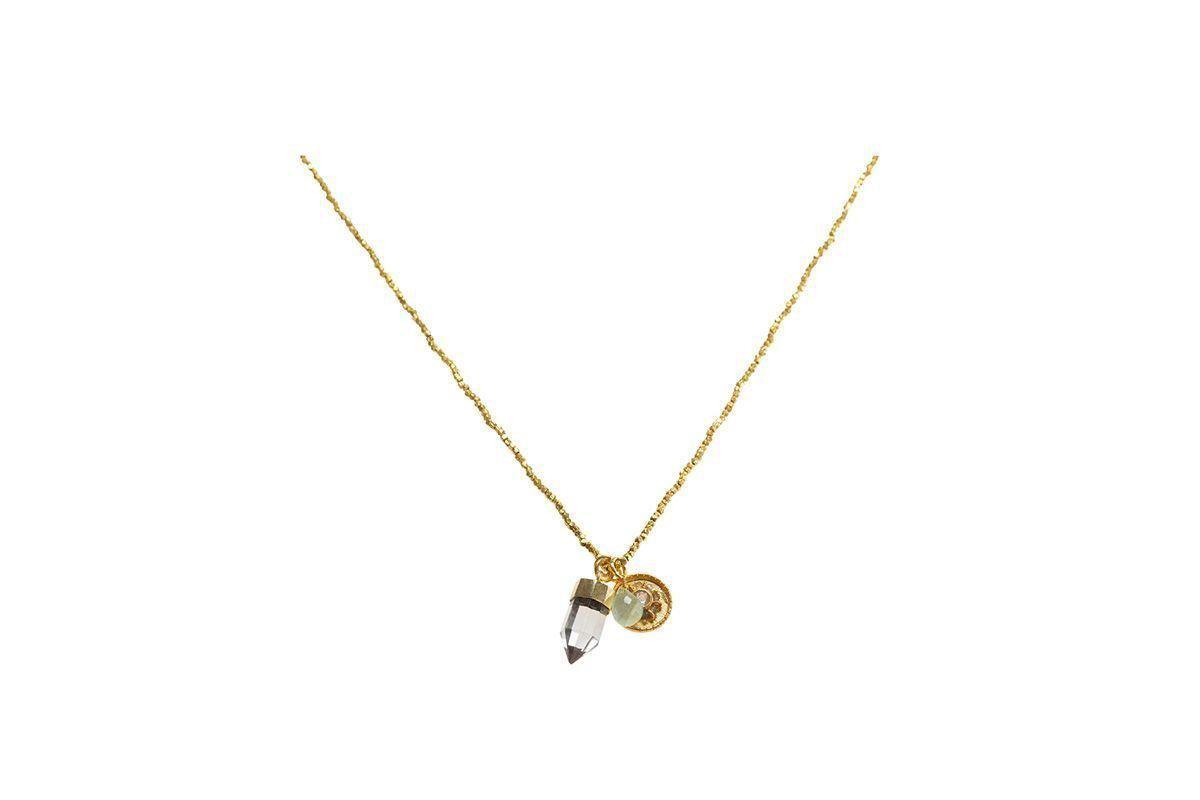 The Pandita Charm Necklace by nkuku features a small clear quartz point with a gold cap and a round engraved gold charm, both suspended together. This unique handmade jewelry piece is shown against a plain white background.