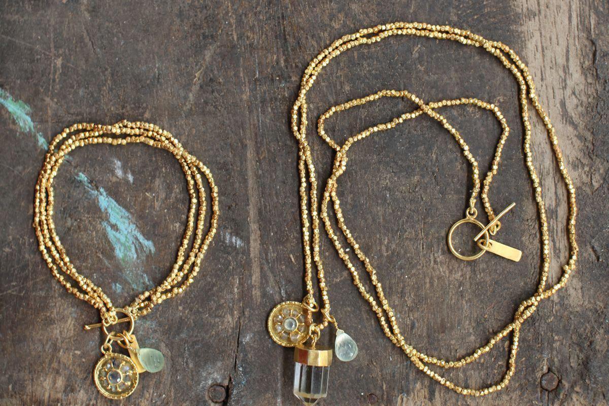 The Pandita Charm Necklace by nkuku sits on a rustic wooden surface, paired with a gold beaded bracelet. Each handmade piece features round medallions and crystal quartz beads, highlighting the collection’s simple yet elegant design.