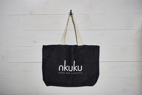 The nkuku Jute & Cotton Tote Bag, a large black handmade-style tote with sturdy beige handles and "nkuku HOME AND LIFESTYLE" in white on the front, hangs on a hook against a white wooden wall.