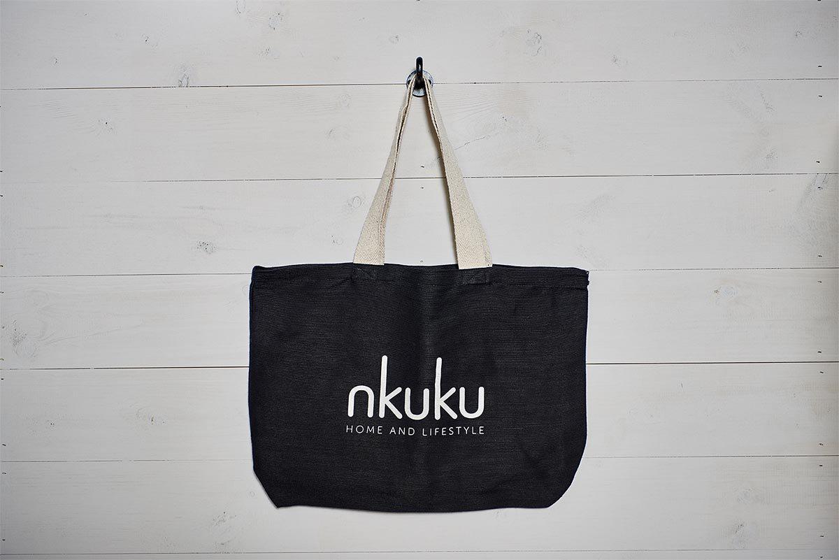 The nkuku Jute & Cotton Tote Bag, a large black handmade-style tote with sturdy beige handles and "nkuku HOME AND LIFESTYLE" in white on the front, hangs on a hook against a white wooden wall.