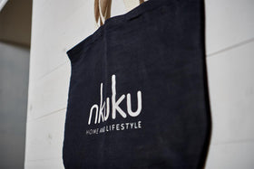 The Jute & Cotton Tote Bag by nkuku, in black with beige handles, hangs on a white wooden wall. The bag displays the white nkuku logo in lowercase, with "HOME AND LIFESTYLE" below it in small uppercase letters.